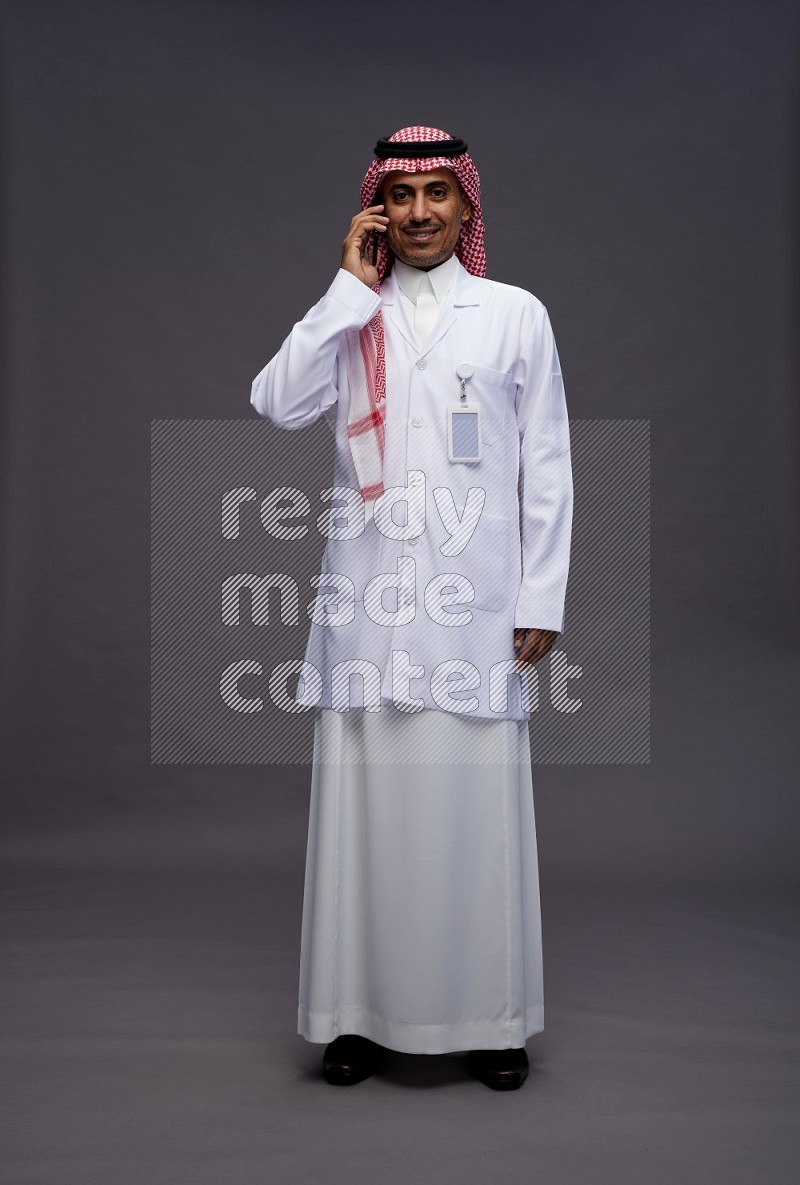 Saudi man wearing thob with lab coat and shomag with pocket employee badge standing talking on phone on gray background