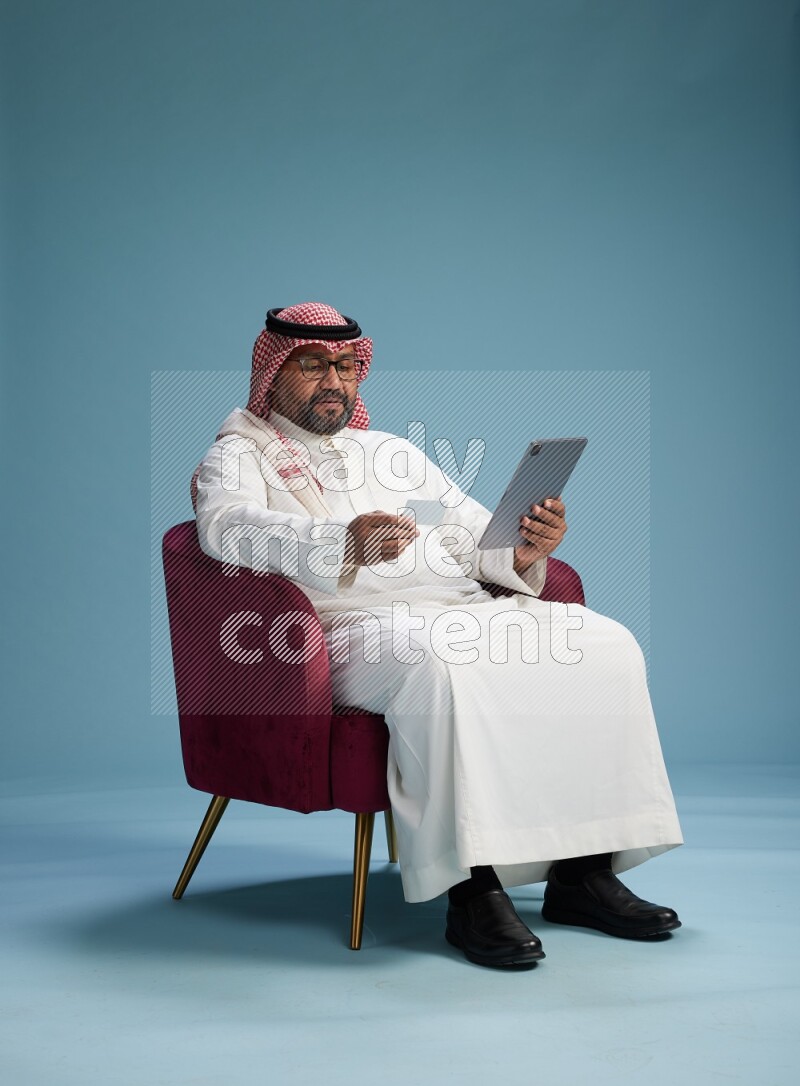 Saudi Man with shimag sitting on chair holding ATM card while working on tablet on blue background