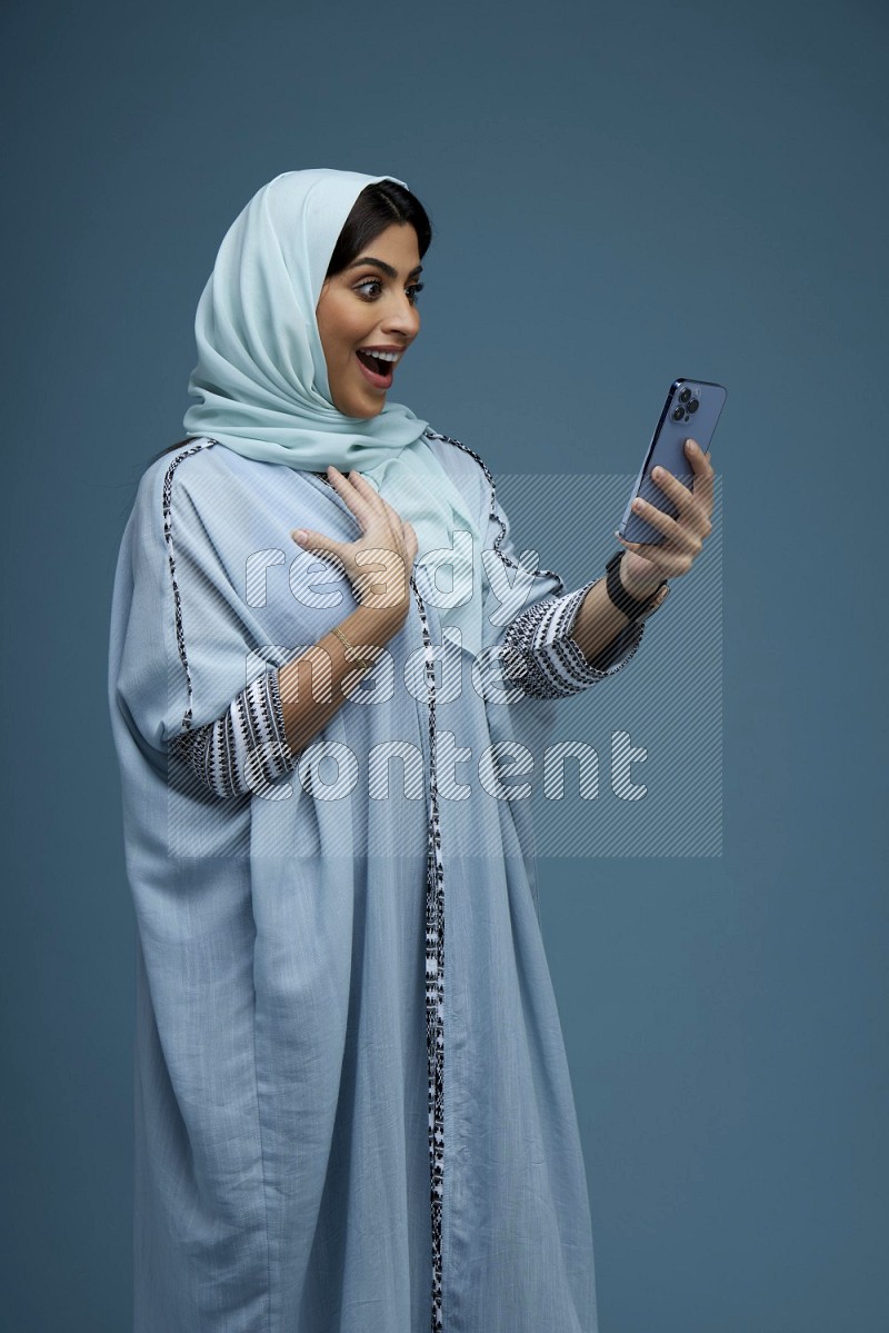 A Saudi woman posing with her phone in a blue background wearing a blue Abaya with hijab