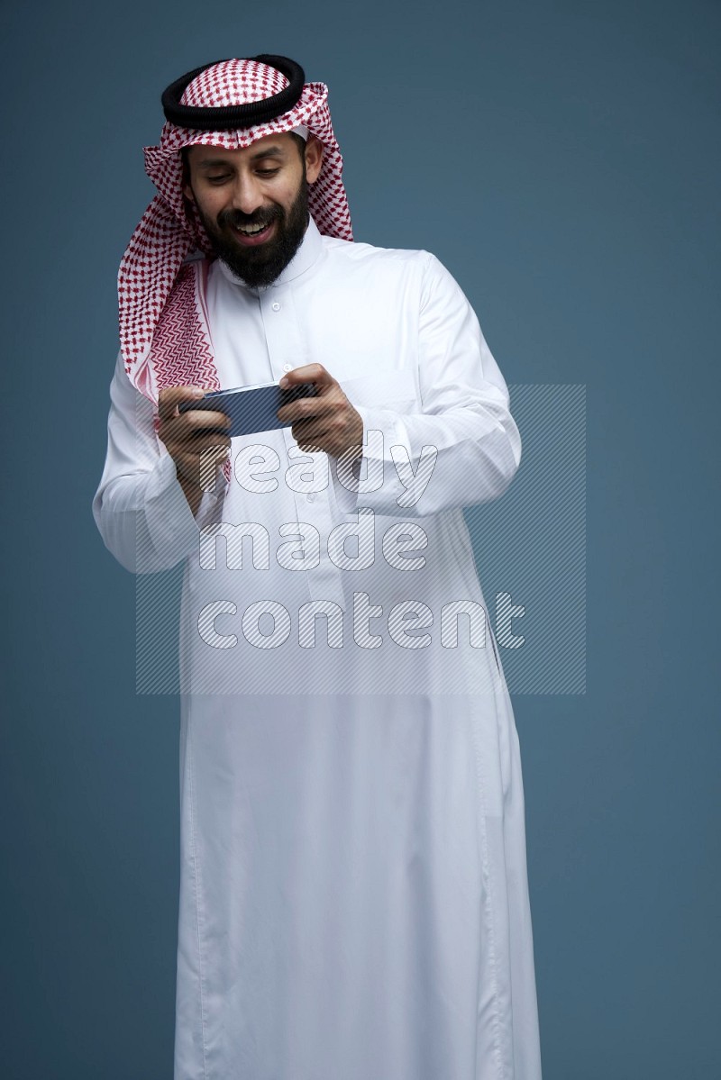 A man Playing a Game on his phone in a blue background wearing Saudi Thob with Shomag