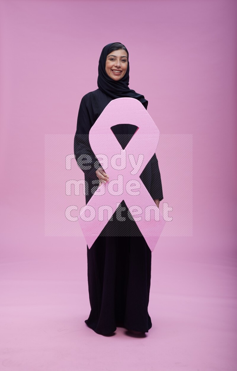 Saudi woman wearing pink ribbon on abaya standing holding awareness ribbon on plain pink background