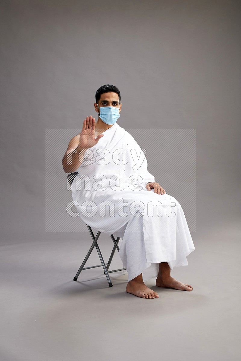 A man wearing Ehram with face mask sitting on chair Interacting with the camera on gray background