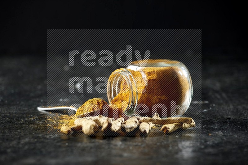 A flipped glass spice jar and metal spoon full of turmeric powder and powder spilled out of it with dried whole fingers on textured black flooring