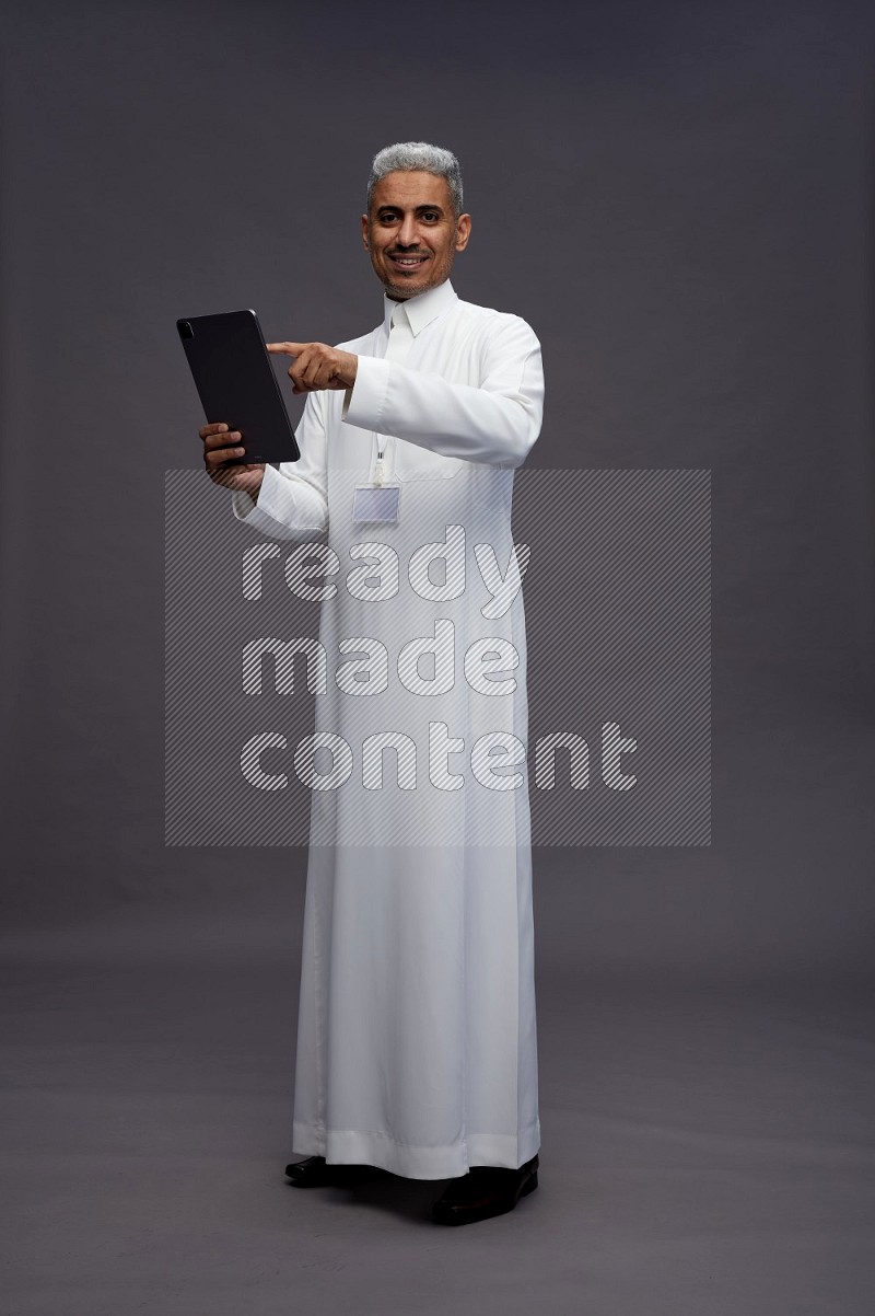 Saudi man wearing thob with neck strap employee badge standing working on tablet on gray background