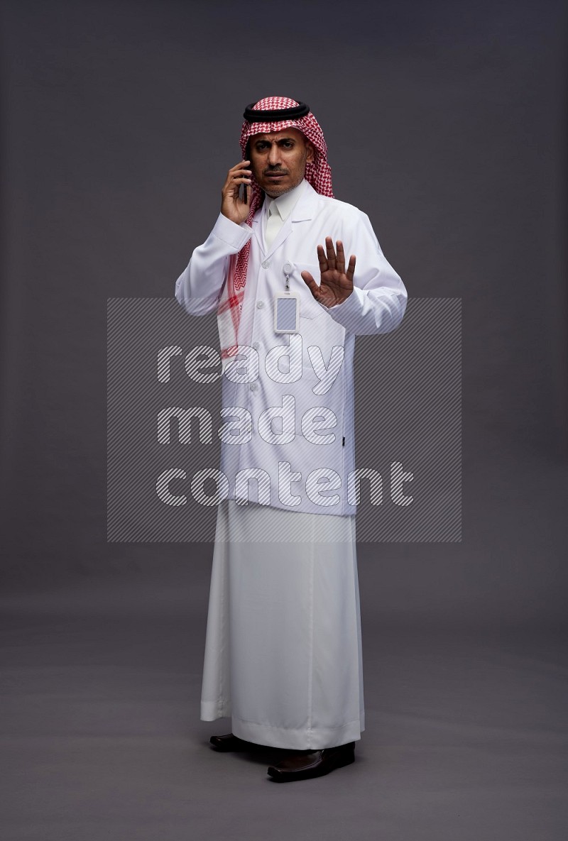 Saudi man wearing thob with lab coat and shomag with pocket employee badge standing talking on phone on gray background