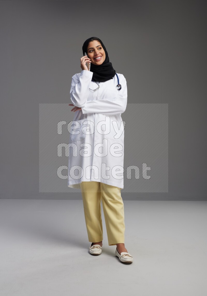 Saudi woman wearing lab coat with stethoscope standing talking on phone on Gray background