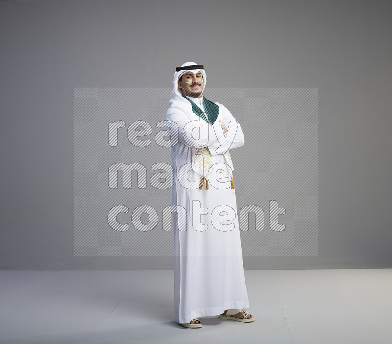 A Saudi man standing wearing thob and white shomag with face painting and Saudi flag scarf on gray background