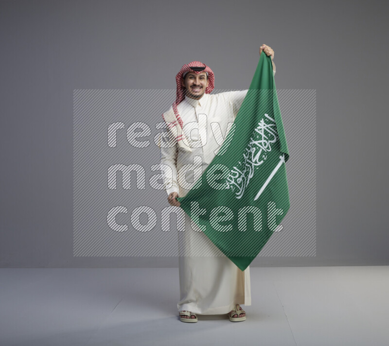 A saudi man standing wearing thob and red shomag holding big saudi flag on gray background