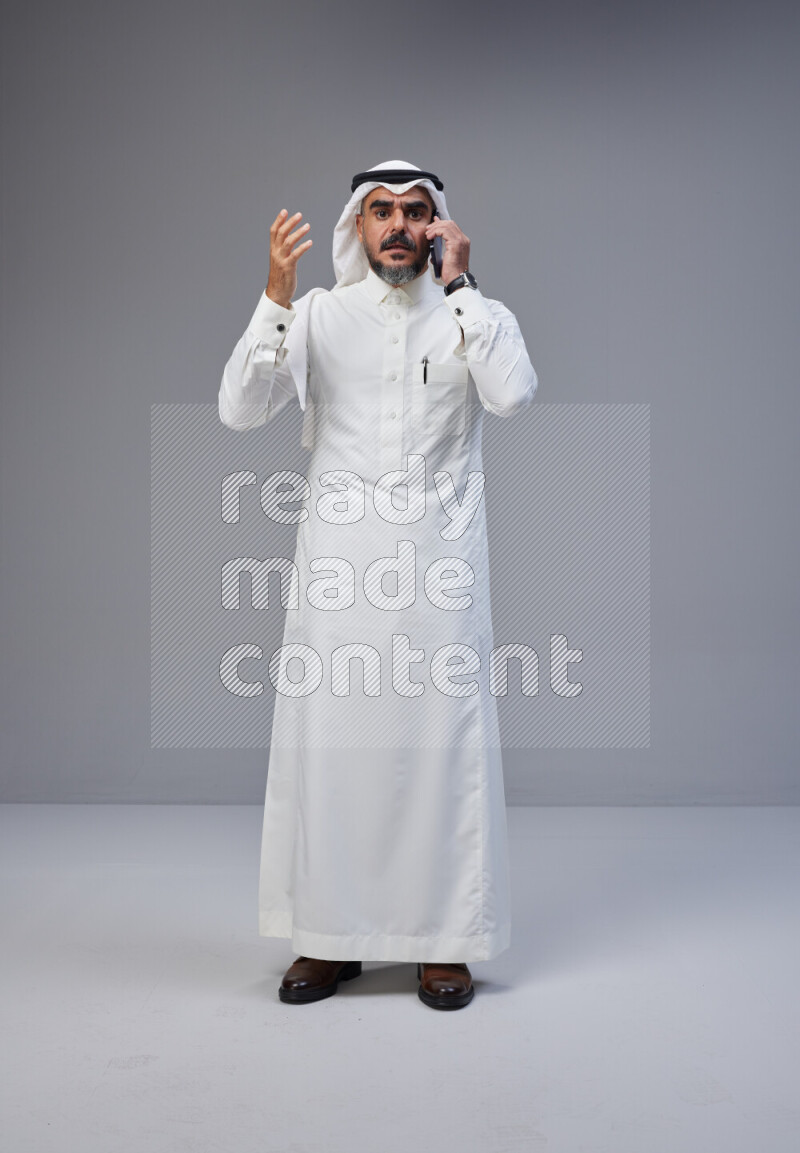 Saudi man Wearing Thob and white Shomag standing talking on phone on Gray background