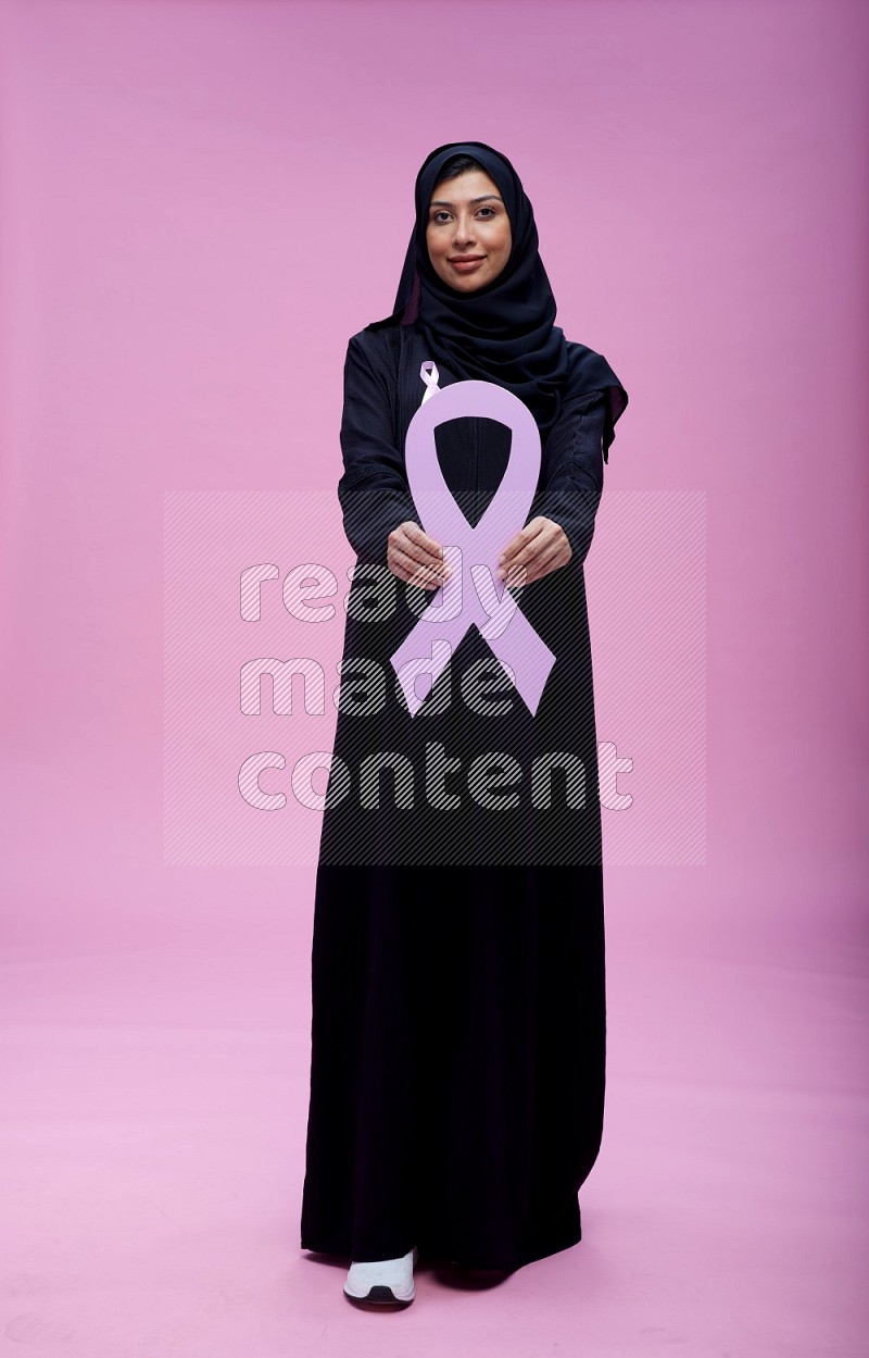 Saudi woman wearing pink ribbon on abaya standing holding awareness ribbon on plain pink background