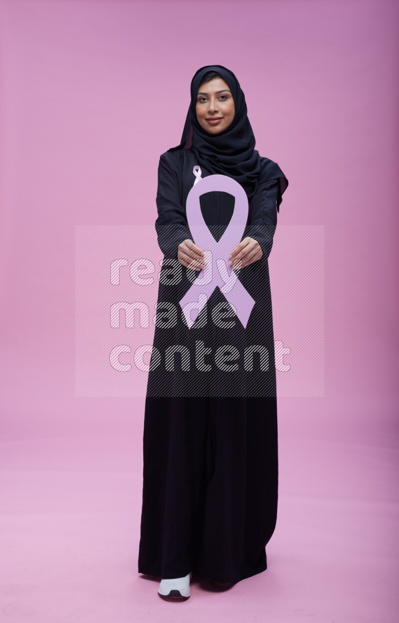 Saudi woman wearing pink ribbon on abaya standing holding awareness ribbon on plain pink background