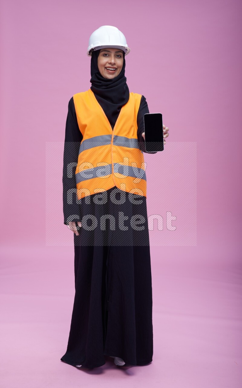 Saudi woman wearing Abaya with engineer vest and helmet standing showing phone to camera on pink background