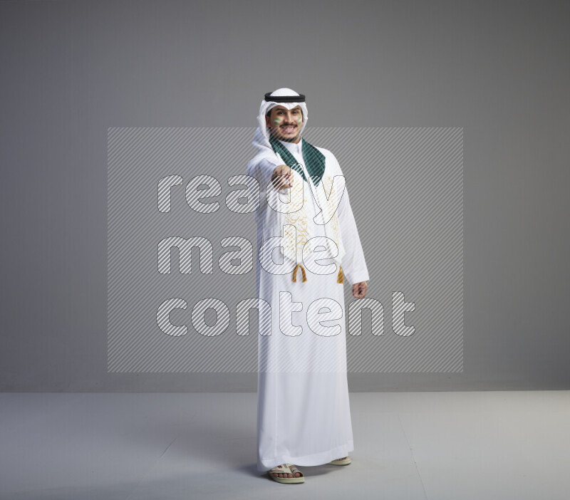 A Saudi man standing wearing thob and white shomag with face painting and Saudi flag scarf on gray background