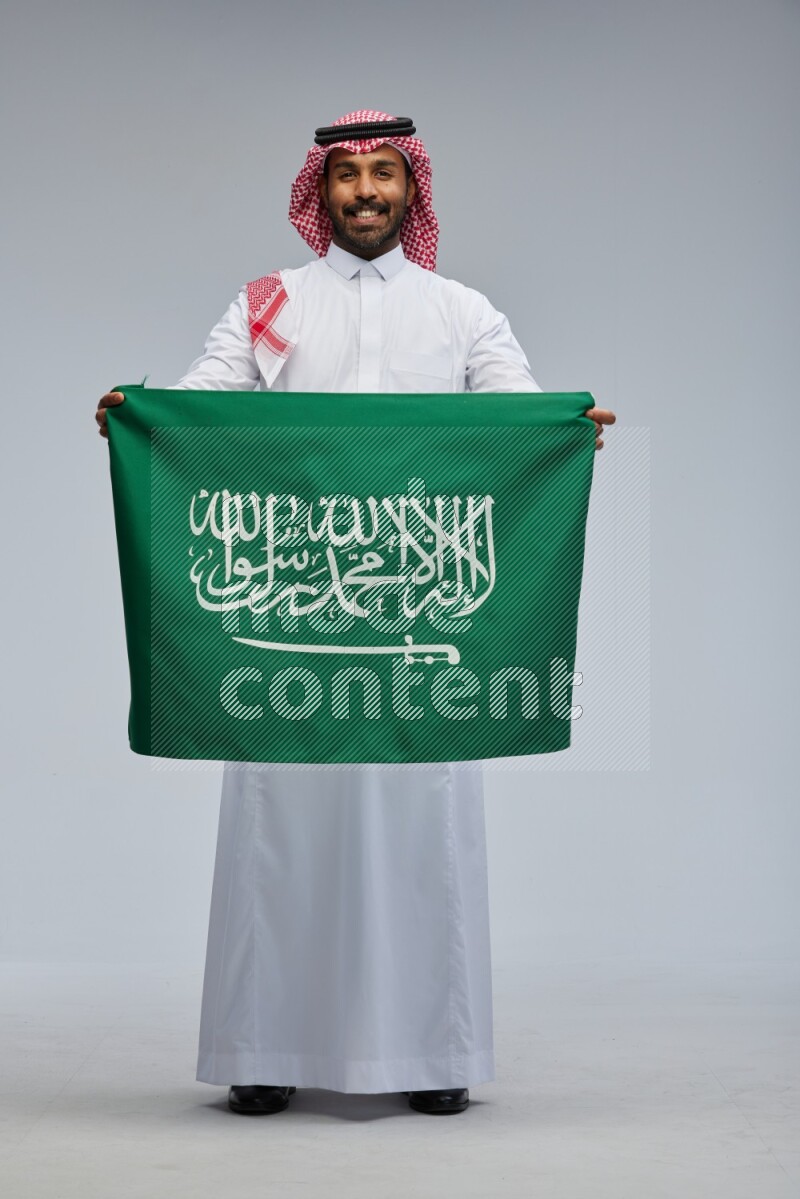 Saudi man Wearing Thob and shomag standing holding Saudi flag on Gray background