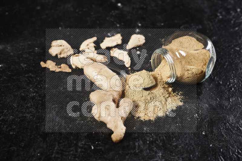 A glass jar full of ground ginger powder flipped with some spilling powder on black background