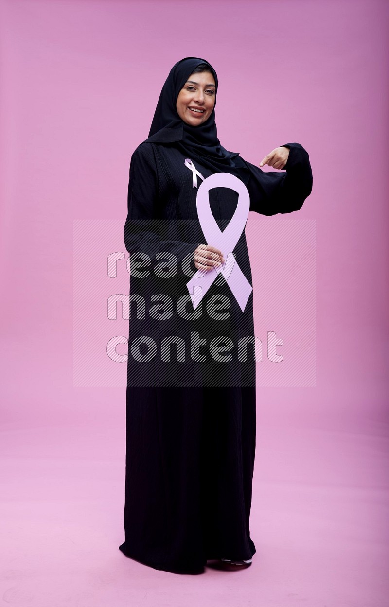 Saudi woman wearing pink ribbon on abaya standing holding awareness ribbon on plain pink background