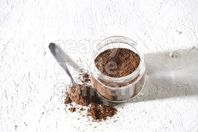 A glass jar full of cloves powder with a metal spoon on a textured white flooring