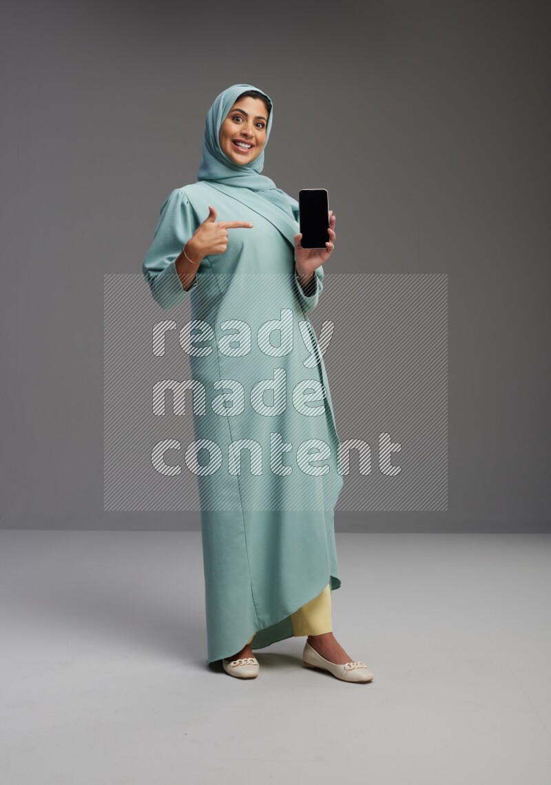 Saudi Woman wearing Abaya standing showing phone to camera on Gray background