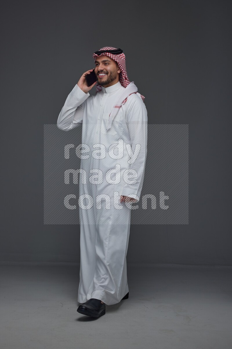 A Saudi man wearing Thobe and Shmagh talking in the phone eye level on a gray background