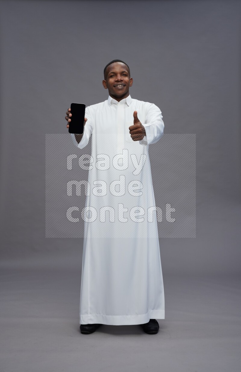 Saudi man wearing thob standing showing phone to camera on gray background