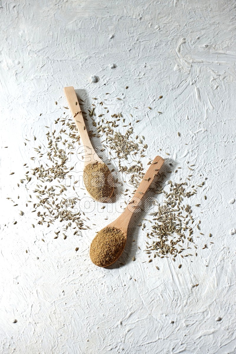 2 Wooden spoons full of cumin powder and cumin seeds on textured white flooring