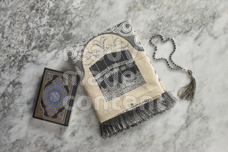 A folded prayer mat with different elements such as quran and prayer beads on grey marble background