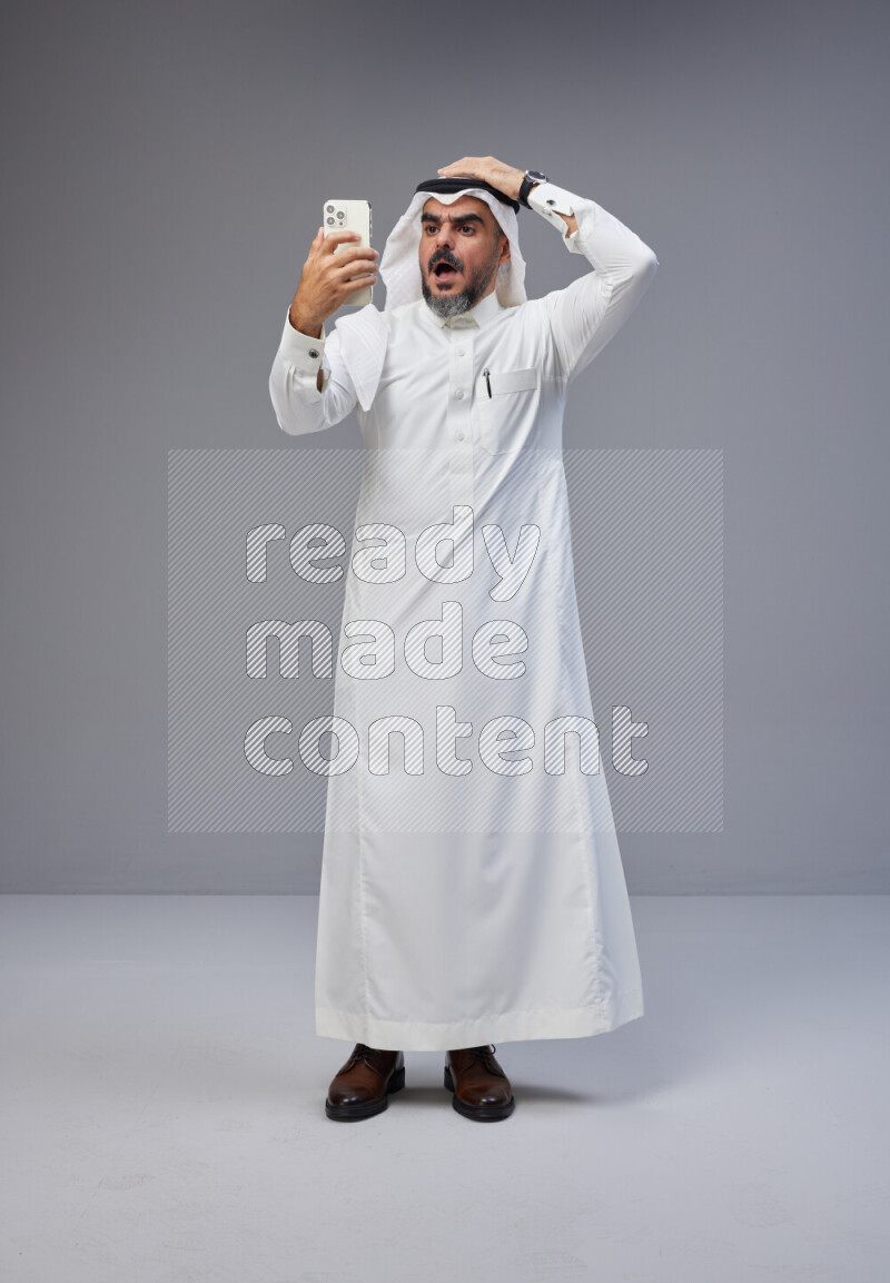Saudi man Wearing Thob and white Shomag standing texting on phone on Gray background
