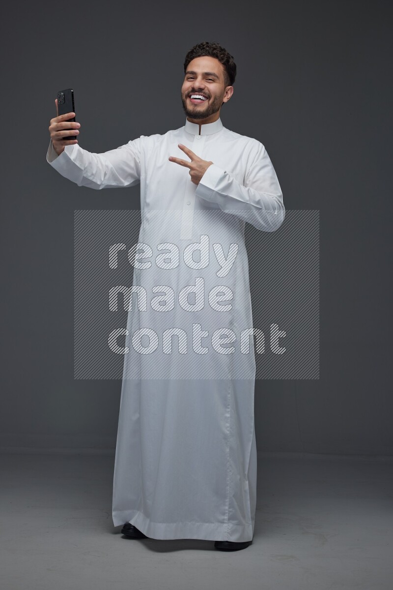 A Saudi man wearing Thobe and taking selfie with his phone making different poses eye level on a gray background