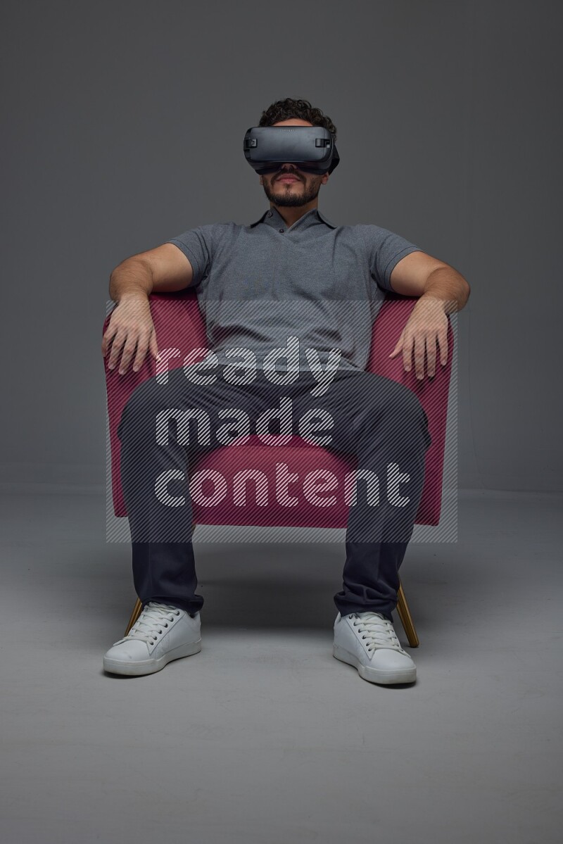 A man wearing casual and wearing VR while sitting on a burgundy chair eye level on a gray background