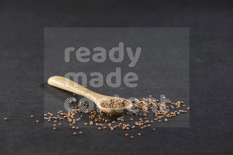 A wooden spoon full of mustard seeds on a black flooring