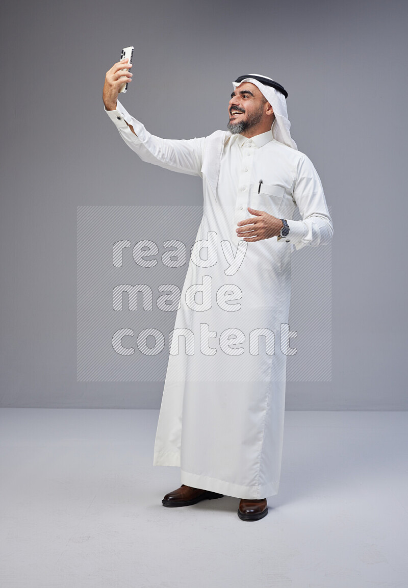 Saudi man Wearing Thob and white Shomag standing taking selfie on Gray background