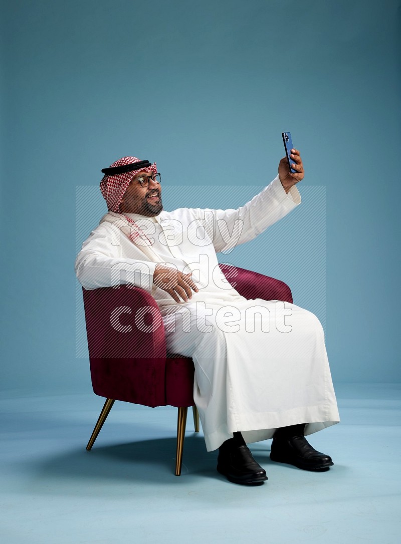 Saudi Man with shimag sitting on chair taking selfie on blue background