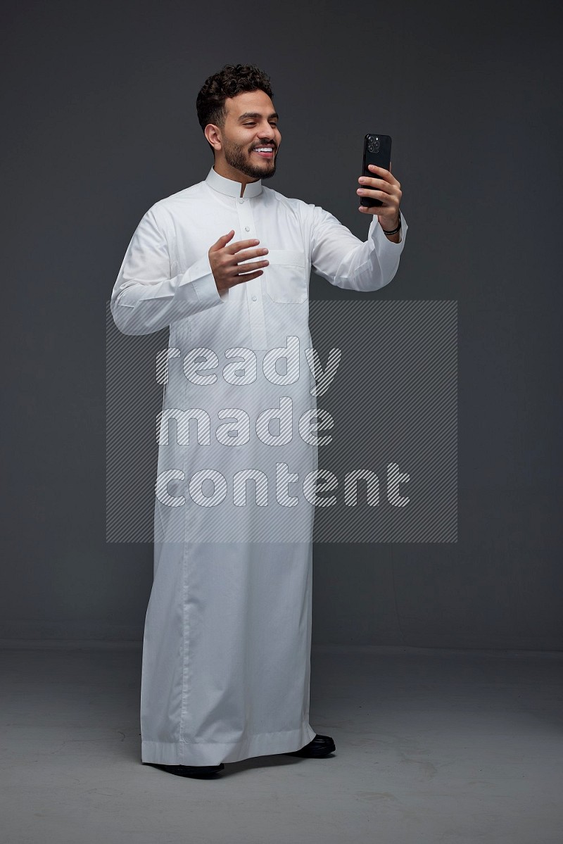 A Saudi man wearing Thobe and taking selfie with his phone making different poses eye level on a gray background