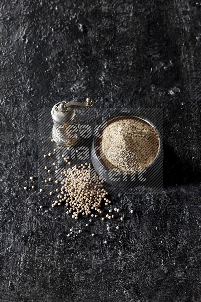 Black pottery bowl full of white pepper powder with pepper beads and metal pepper grinder on textured black flooring