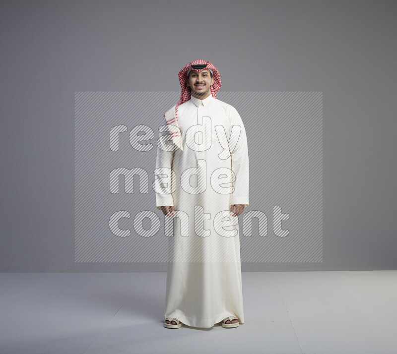 A Saudi man standing wearing thob and red shomag interacting with the camera on gray background