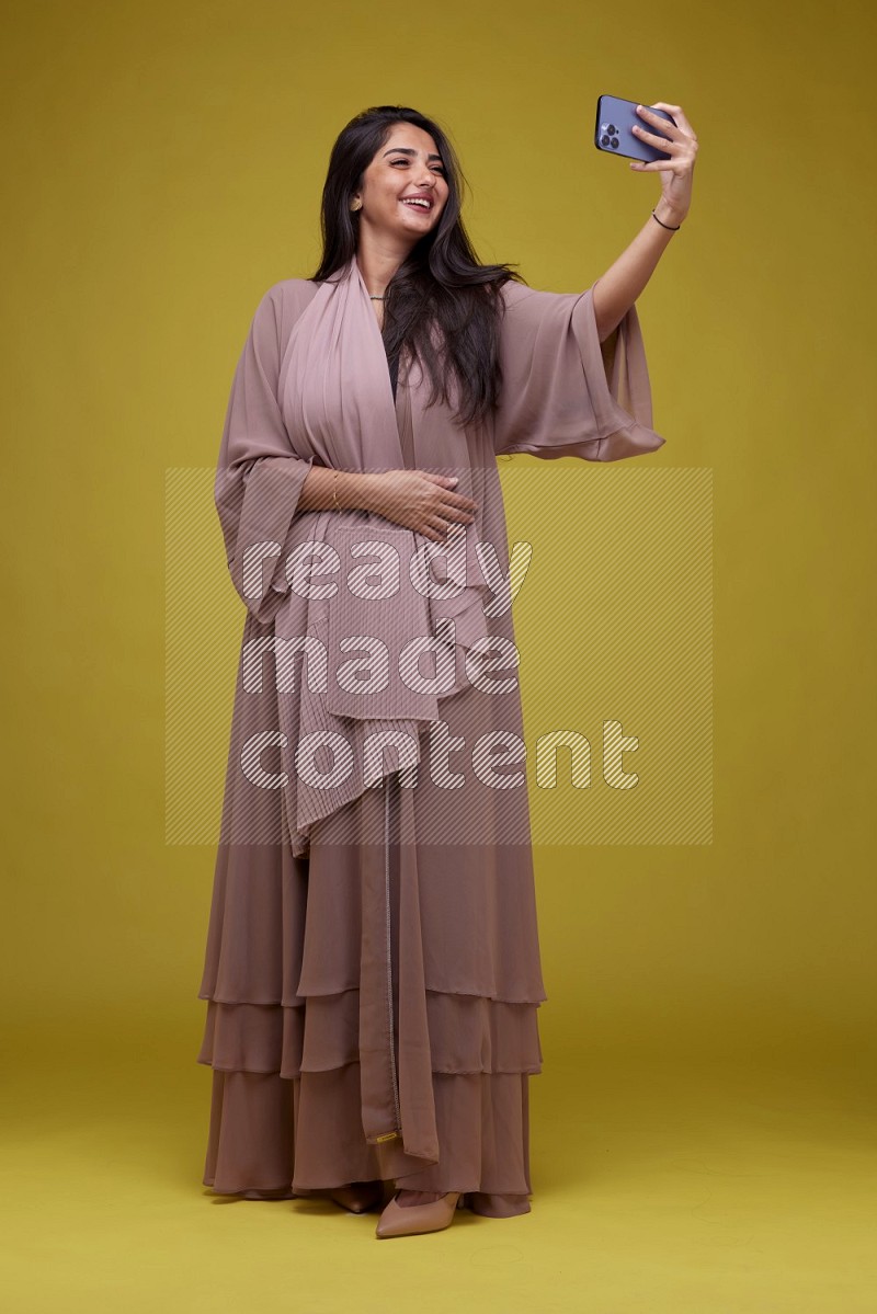 A woman Taking a Selfie on a Yellow Background wearing Brown Abaya