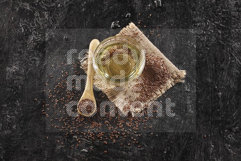 A glass bowl full of flaxseeds oil and wooden spoon full of flaxseeds with seeds spread on burlap fabric on a textured black flooring