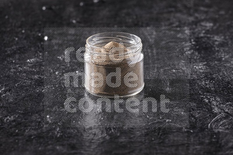 A glass jar full of black pepper powder on a textured black flooring