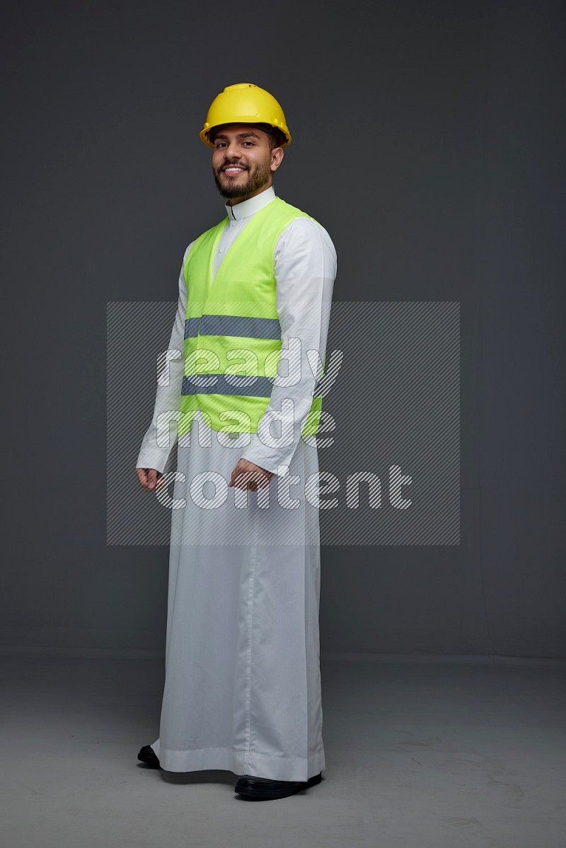 A Saudi man wearing Thobe with a yellow safety vest and white helmet standing and crossing his hands eye level on a gray background