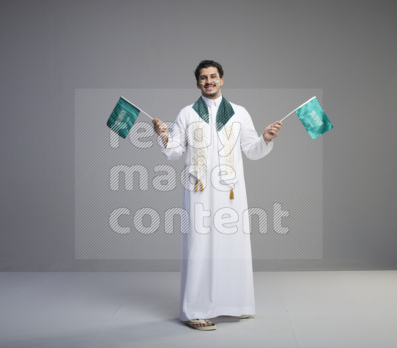 A Saudi man standing wearing thob and saudi flag scarf with face painting holding small Saudi flag on gray background