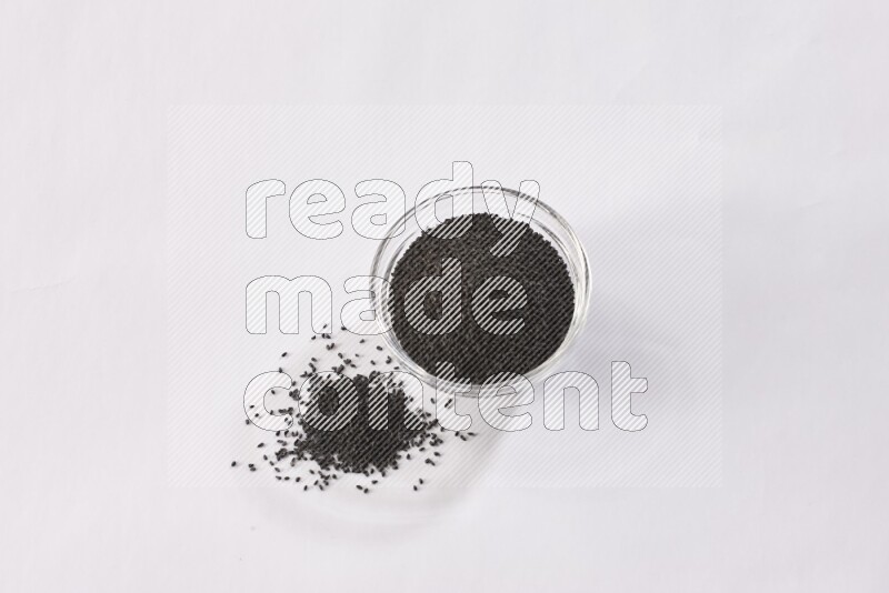 A glass bowl full of black seeds and some more seeds spread next to it on a textured white flooring