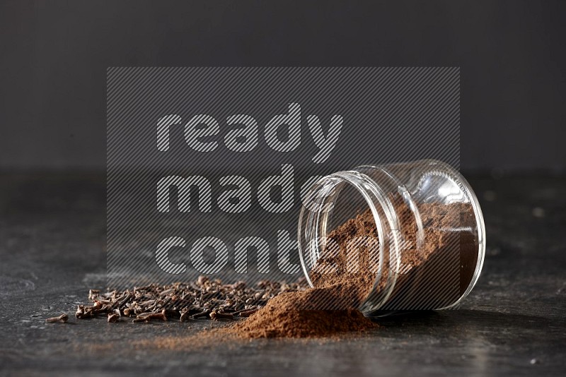 A flipped glass jar full of cloves powder with cloves spread on a textured black flooring