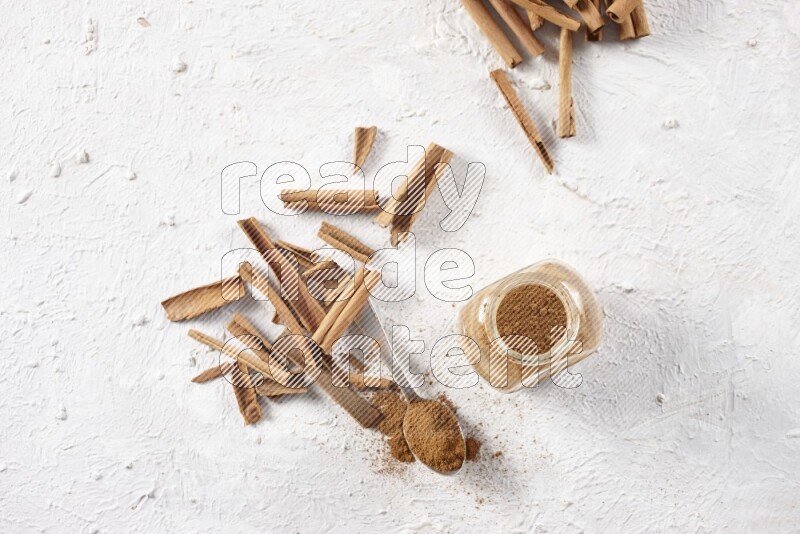 Herbal glass jar full cinnamon powder and a metal spoon surrounded by cinnamon sticks on a white background