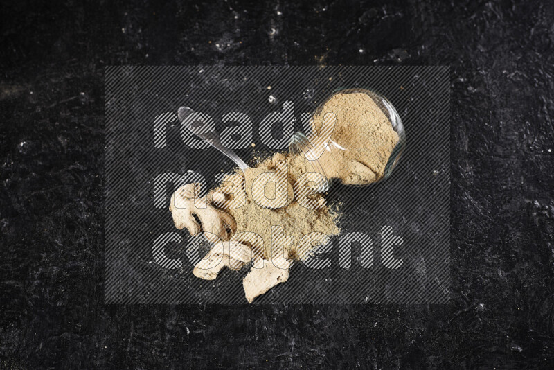 A glass jar full of ground ginger powder flipped with some spilling powder on black background