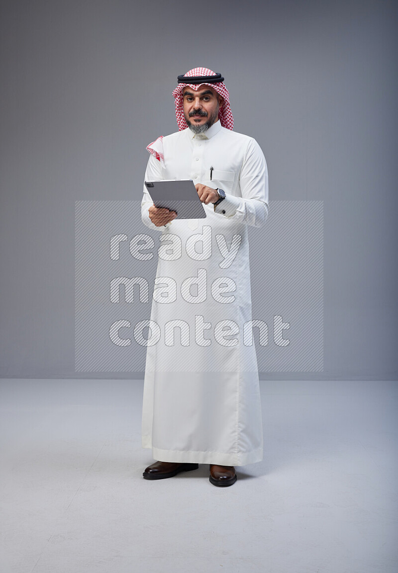 Saudi man Wearing Thob and red Shomag standing working on tablet on Gray background