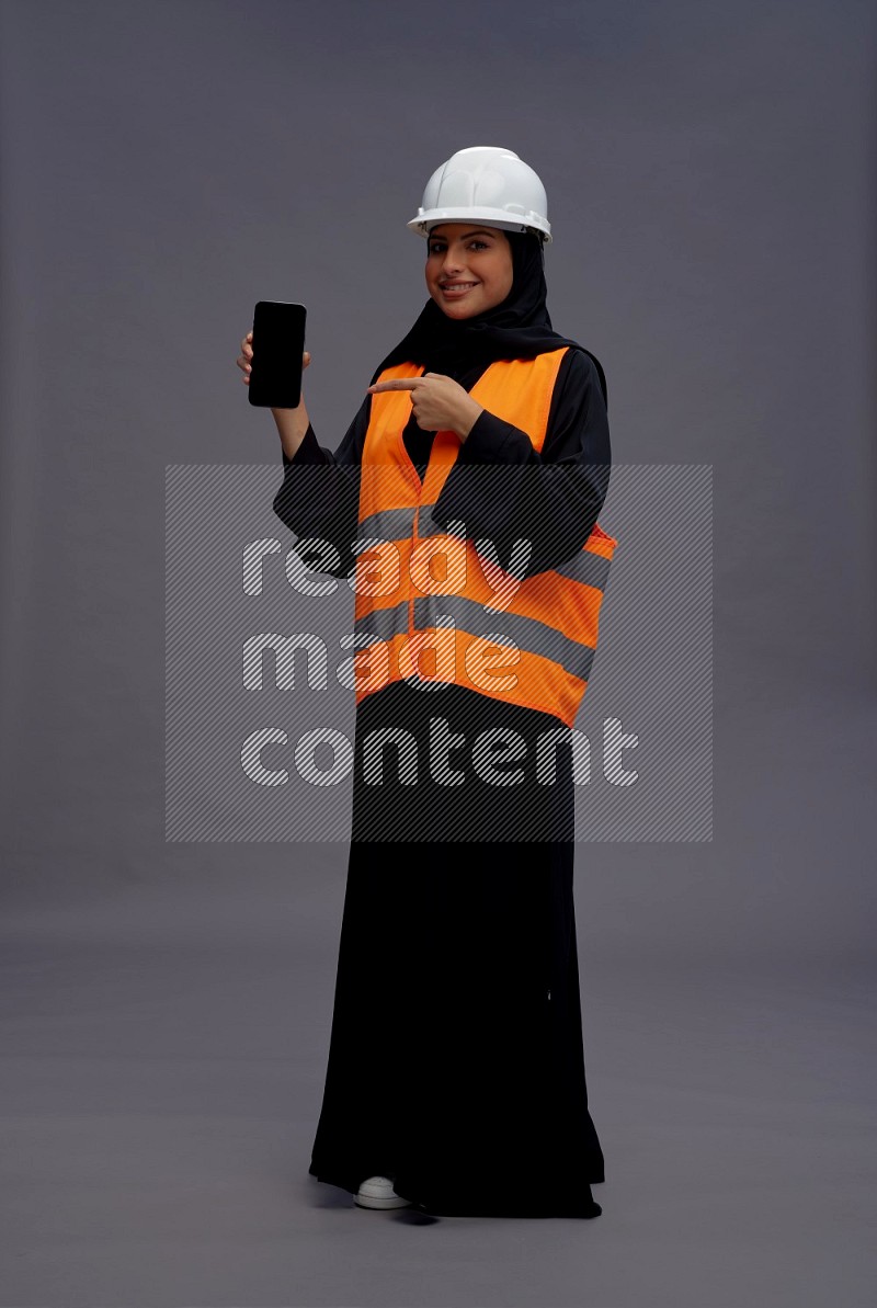 Saudi woman wearing Abaya with engineer vest standing showing phone to camera on gray background