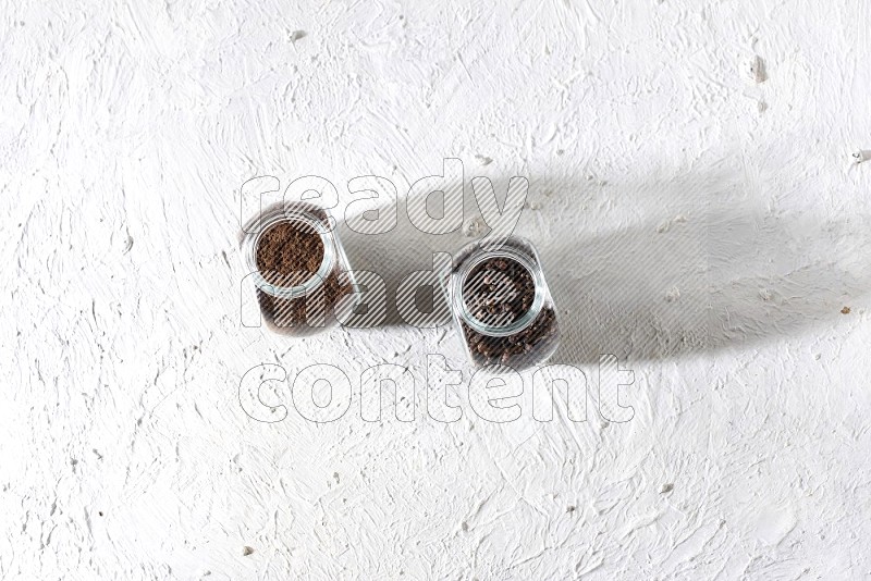 2 glass jars full of cloves powder and cloves grains on a textured white flooring