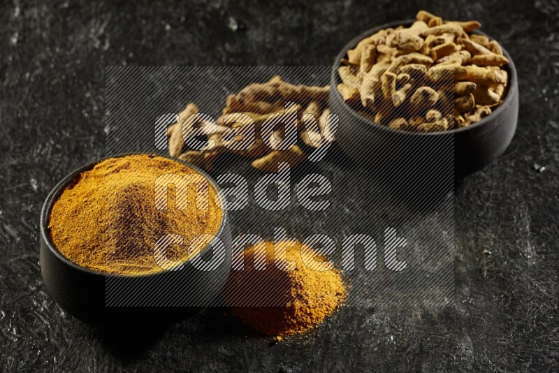 2 pottery black bowls, one full of turmeric powder and the other full of dried turmeric whole fingers on a textured black flooring