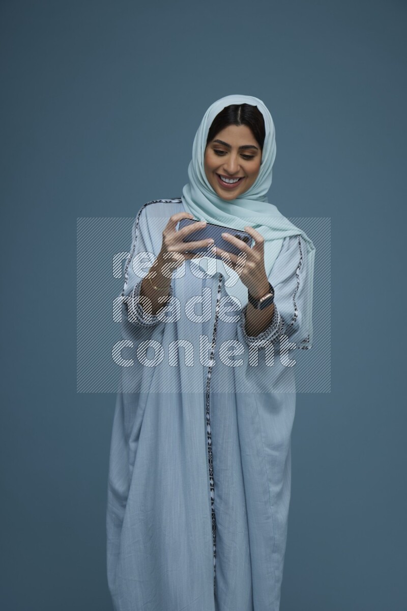 A Saudi woman Playing a Game on her phone on a blue background wearing a blue Abaya with hijab