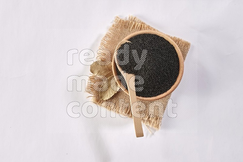 A wooden bowl and wooden spoon full of black seeds on a piece of burlap on a white flooring in different angles